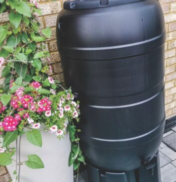 A black water butt sitting next to a pot of flowers.