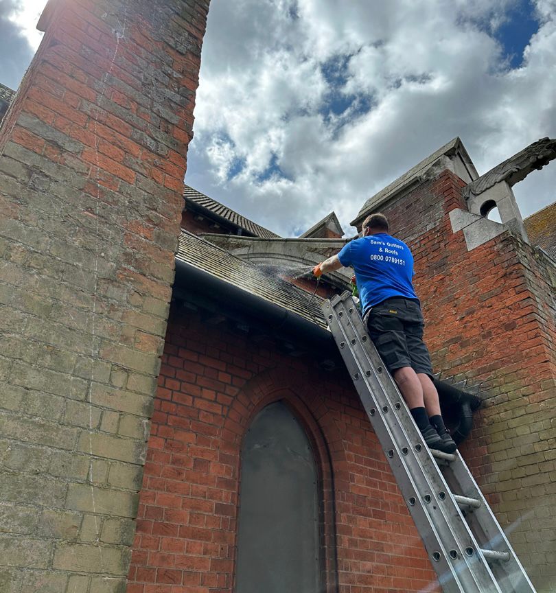 A roofer stood at the top of a ladder spraying the roof of a commercial building after cleaning it.