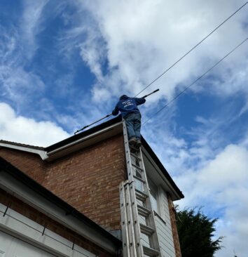 Looking up at a roofer standing on the top of a ladder clearing gutters using an extendable pole.